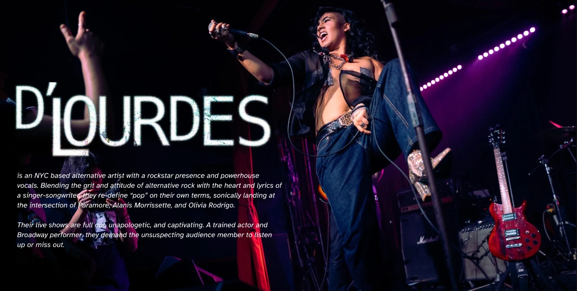 D&rsquo;LOURDES mid-performance on stage, microphone raised, in front of stage lights.