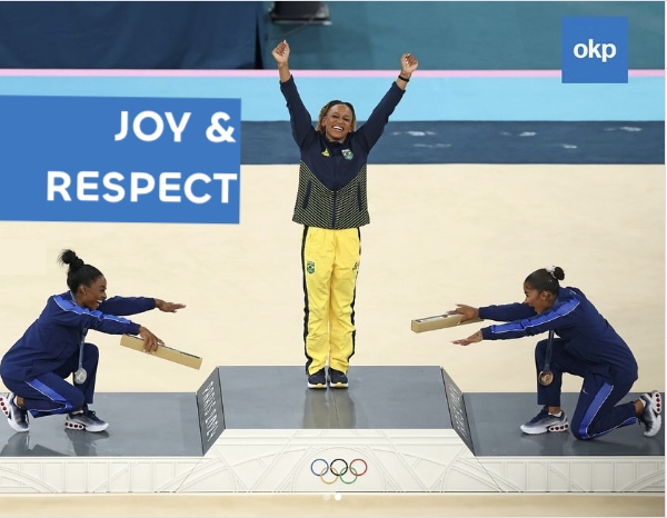 Rebecca Andrade of Brazil stands on the gold medal step of the floor podium at the 2024 Paris Olympics. Jordan Chiles and Simone Biles bow to her from either side.