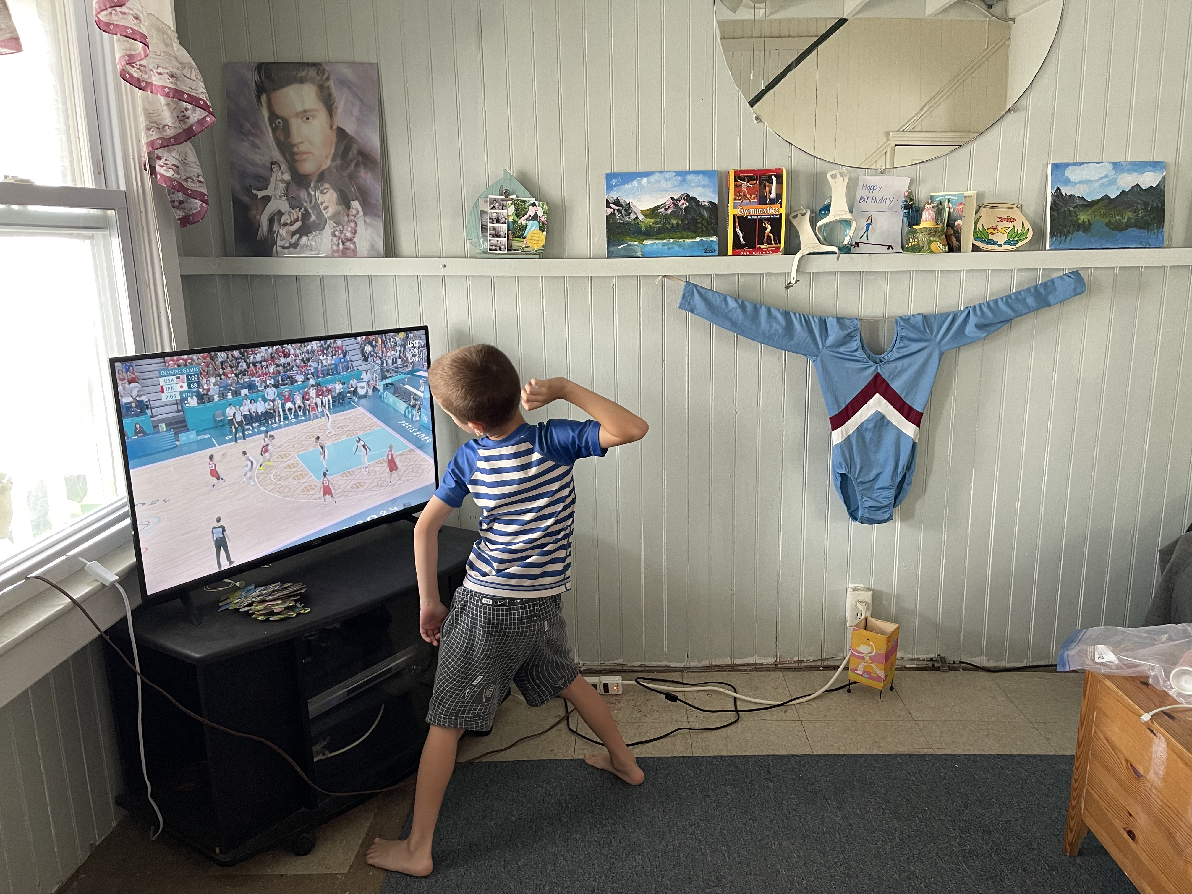A kid watches Olympic basketball on TV next to Wendy Jo's gymnastics shrine — a blue childhood leotard hanging on the wall, a Gymnastics book and other mementos on the shelf.